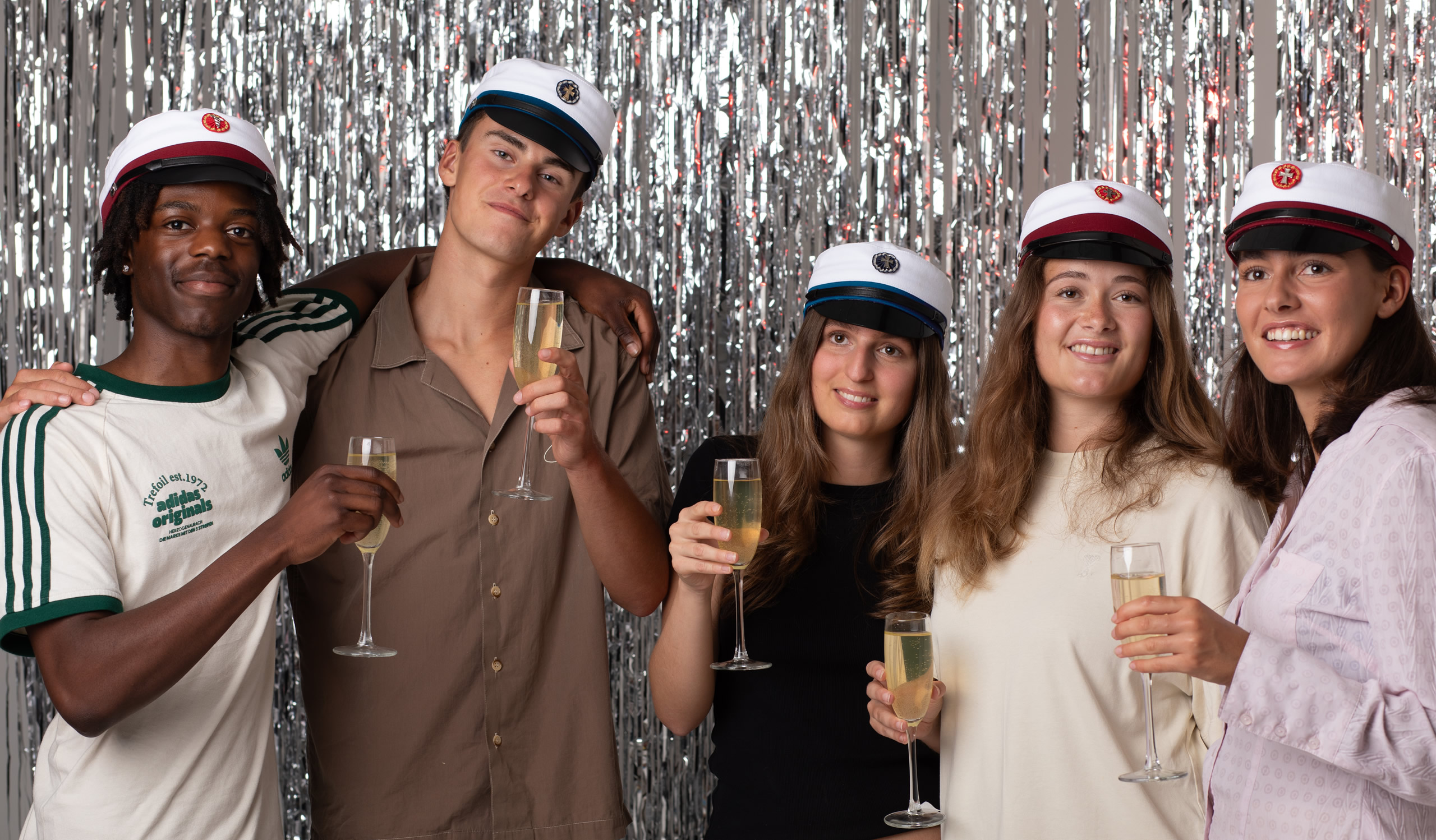 5 glade studenter som har fået der studenterhue på som fejer det med champagne til en baggrund med sølvskimler på en hvid væg